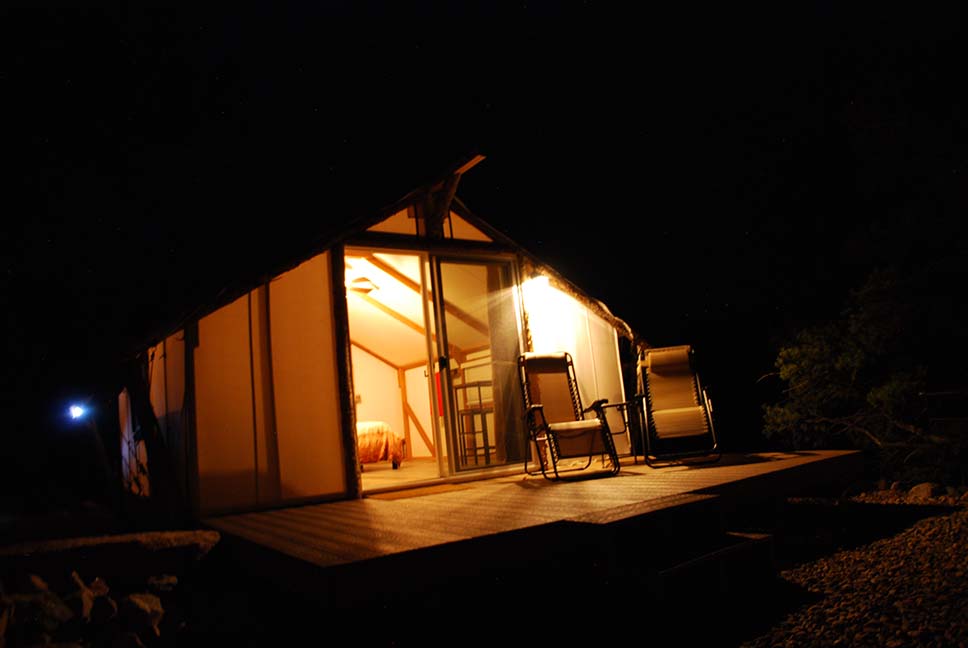 view of a glamping tent at night. the fire is blazing in the firepit as the light from within the tent gently invites you in